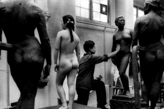 A naked female model poses in front of a sculpture.