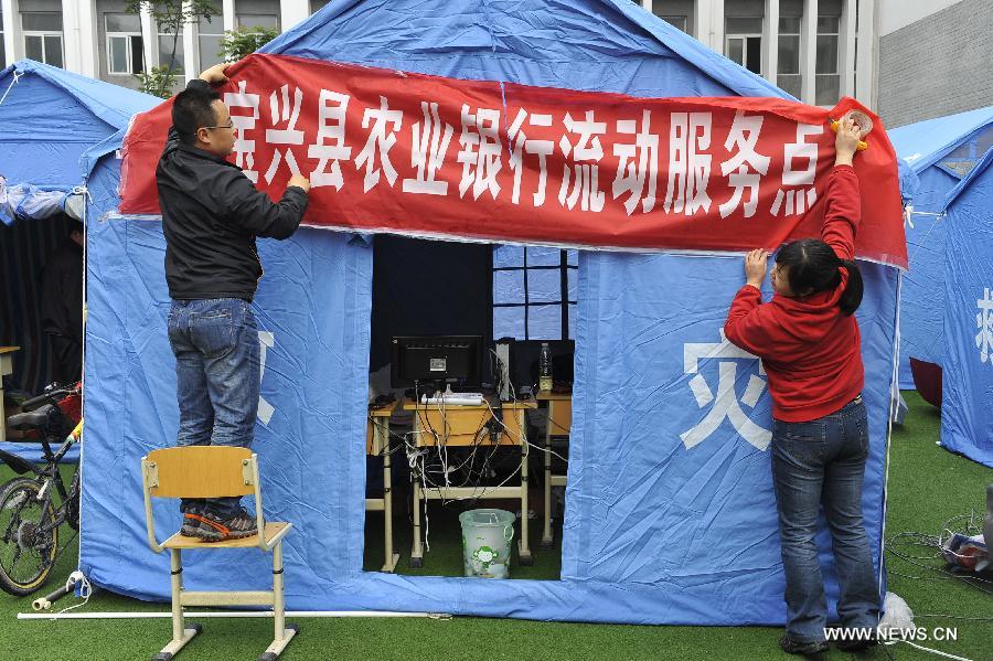Staff members hang a banner on the makeshift office of Agriculture Bank of China at a school served as an evacuation settlement for displaced people in the quake-hit Baoxing County, southwest China's Sichuan Province, May 2, 2013. Some 4,000 displaced people live the settlement, the largest in the county, after a strong earthquake hit Baoxing last month. At present, basic living needs of those people could be guaranteed in terms of food, drink, healthcare service and power supply. (Xinhua/Lu Peng)