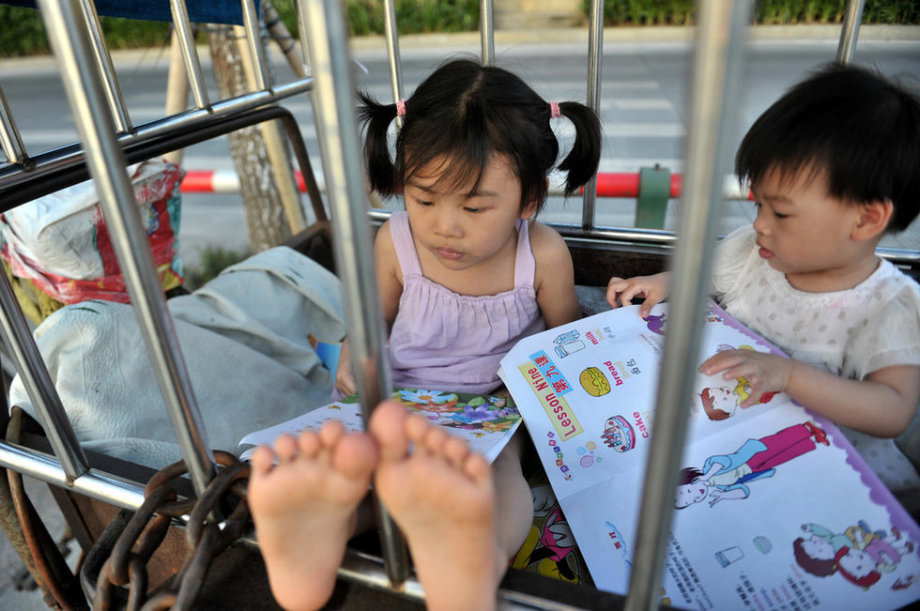 The two girls read a book in a cage-like tricycle, waiting for their grandmother who does laundry by the river in S China’s Guangxi, June 18, 2013. (Photo/ youth.cn)